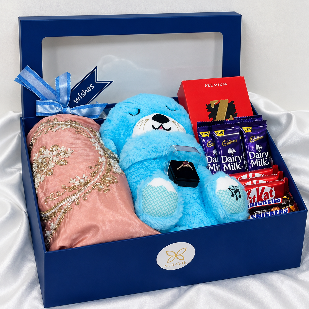 Luxury women’s gift hamper with blue breathing teddy, embroidered pink dress, chocolates, jewellery ring and premium gift box.
