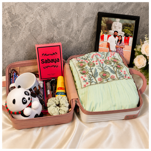 Premium women’s suitcase gift hamper with western dress, photo frame, perfume, panda soft toy, coffee mug, chocolates, sunscreen, eyeliner, lipstick and hair accessories.